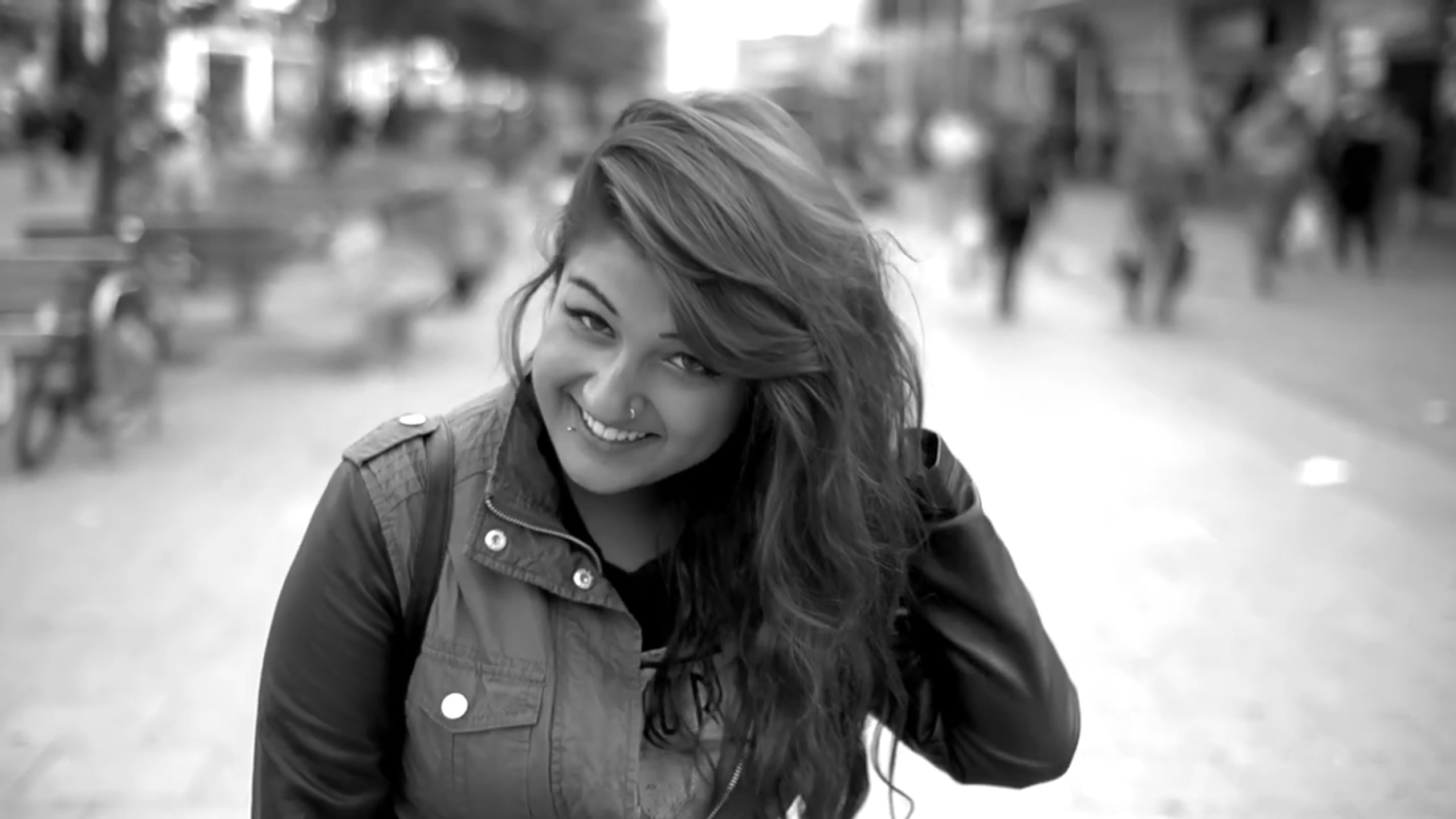 A woman adjusting her hair in the street, looking sheepishly at the camera in black and white.